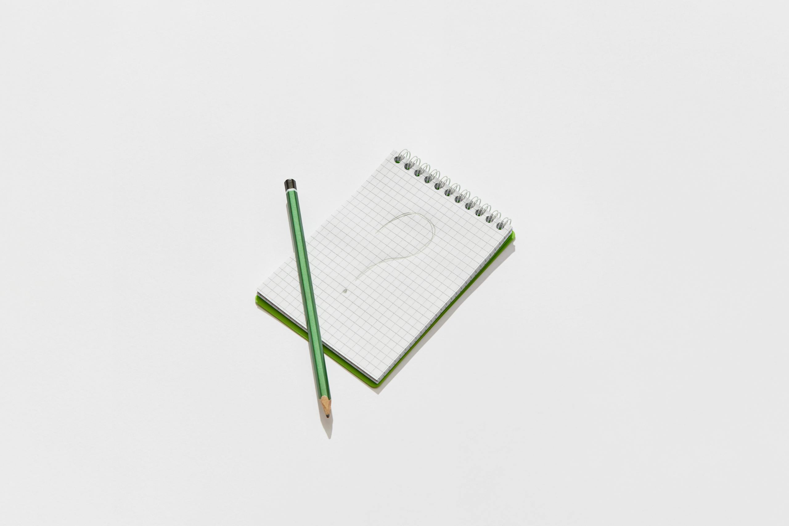 Minimalist photo of a green notepad and pencil on a white background with a drawn question mark.